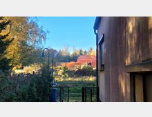 Blick zwischen einem Gebäude mit brauner Fassade und einer Hecke hindurch auf ein Grundstück mit einem Zaun, Rasenfläche, Bäumen und roten Ziegeldächern im Hintergrund. Im oberen Bereich sind blauer Himmel und Wald sichtbar.