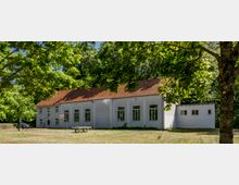 Weiße, einstöckige Gebäude mit roten Ziegeldächern, umgeben von Bäumen und einer kleinen Wiese im Vordergrund; links ein Basketballkorb und eine Sitzbank, rechts ein seitlicher Anbau.