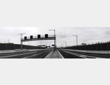 Eine vierspurige Autobahn mit getrennten Fahrbahnen, flankiert von hohen Lärmschutzwänden. Im Vordergrund befinden sich Verkehrsschilder und eine digitale Anzeigetafel auf einer Metallbrücke, der Himmel ist wolkig und grau.