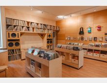 Interior of a modern record shop with light wood floors and walls, featuring built-in shelving filled with vinyl records and large speaker cabinets. Low display bins of records sit in the center, and a pegboard wall holds small merchandise and magazines under track lighting.