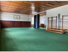 Ein Sportraum mit grünem Teppichboden, einer Holzverkleidung an der unteren Wandhälfte und einer Holzdecke mit eingelassenen Leuchten. An der rechten Wand befinden sich Klettergerüste, Hanteln und gestapelte Bänke, während an der Stirnwand ein Basketballkorb montiert ist.