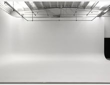 Leerer Fotostudio-Raum mit einer weißen, nahtlosen Hintergrundwand, einer hohen Decke mit freiliegenden Metallrohren und Beleuchtung. Der Boden ist glatt und der Raum wirkt minimalistisch.