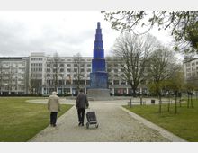 Städtischer Platz mit einem blauen, mehrstufigen Glasturm in der Mitte, umgeben von Bäumen und Grünflächen; im Hintergrund stehen moderne mehrstöckige Gebäude. Zwei Personen gehen auf einem gepflasterten Weg in Richtung der Skulptur.