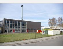 Industriegebäude mit großer Glasfassade und einem hohen Schornstein, umgeben von einer Rasenfläche und einem Maschendrahtzaun. Im Vordergrund befindet sich ein kleiner weißer Technikbereich mit gelben Rohrleitungen sowie ein orangefarbener Container.