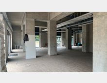 Großer, offener Innenraum eines Rohbaus mit nackten Betonwänden und -säulen, unbehandeltem Fußboden und sichtbaren Lüftungsrohren an der Decke. Durch mehrere bodentiefe Fenster fällt Tageslicht in den Raum, und draußen ist eine Grünfläche sowie ein geparktes Auto sichtbar.