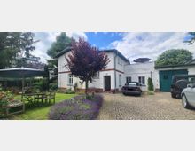 Ein zweistöckiges Wohnhaus mit weißer Fassade und Ziegelakzenten, umgeben von einem gepflegten Garten mit blühenden Lavendelbeeten, einem zentralen Baum und Sitzmöbeln unter einem Sonnenschirm. Rechts im Bild befindet sich ein gepflasterter Hof mit geparkten Autos und einer grünen Garagentür.