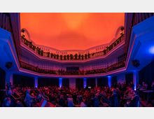 Innenraum eines Veranstaltungsraums mit zwei umlaufenden Emporen und einer kuppelförmigen, orange beleuchteten Decke. Im unteren Bereich sitzt ein Publikum, während weitere Personen auf den Emporen stehen; blaues Licht illuminiert die Wände.