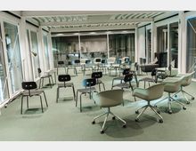 Ein moderner, heller Konferenzraum mit großen Glasfenstern, die den Raum umgeben. Die Bestuhlung besteht aus einem lockeren Kreis aus schwarzen und weißen Stühlen auf einem grünen Teppichboden, ergänzt durch einige Armlehnstühle auf Rollen.