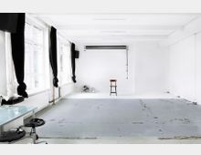 Heller, minimalistisch eingerichteter Raum mit großen Fenstern, schwarzen Vorhängen und weiß gestrichenen Wänden; zentral steht ein einzelner Holzstuhl, im Hintergrund sind zwei Ventilatoren und eine herabhängende Fotostudiorolle sichtbar. Der Boden ist aus grauem Beton mit Abnutzungsspuren.