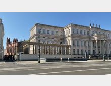 Historisches Gebäude mit klassizistischer Fassade, Säulenreihe und aufwendigen Verzierungen; daneben eine neugotische Kirche aus rotem Backstein. Im Vordergrund Straße mit Zebrastreifen und Gehweg, auf dem einige Personen gehen.