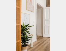 Helles, modern eingerichtetes Zimmer mit Holzfußboden, einer Zimmerpflanze in einem Korb und einer Keramikvase mit Dekozweigen. An der weißen Wand hängen minimalistische Bilder in goldenen Rahmen, im Hintergrund ist eine geöffnete Zimmertür mit Verzierungen sichtbar.