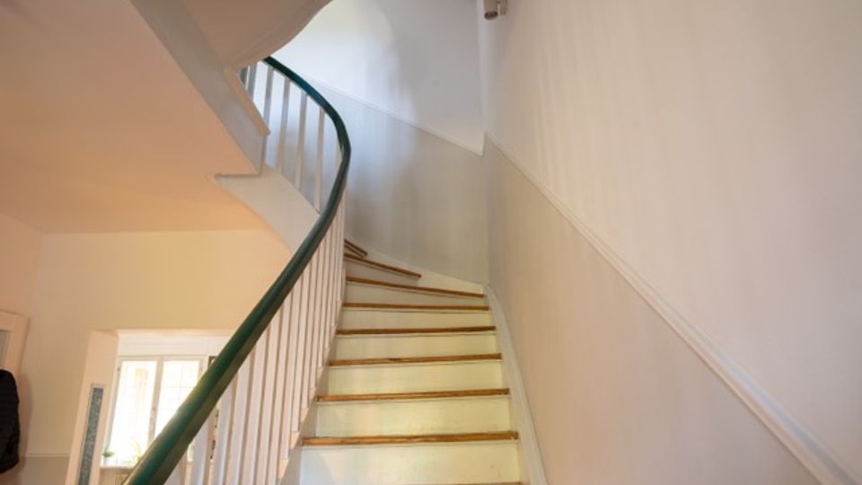 Innenansicht eines Treppenhauses mit einer schmalen, nach oben führenden Holztreppe und einem geschwungenen Geländer mit dunklem Handlauf und weißen Stäben. Weiße Wände und eine Deckenleuchte prägen den hellen Flur, unten ist ein Türdurchgang zu sehen.