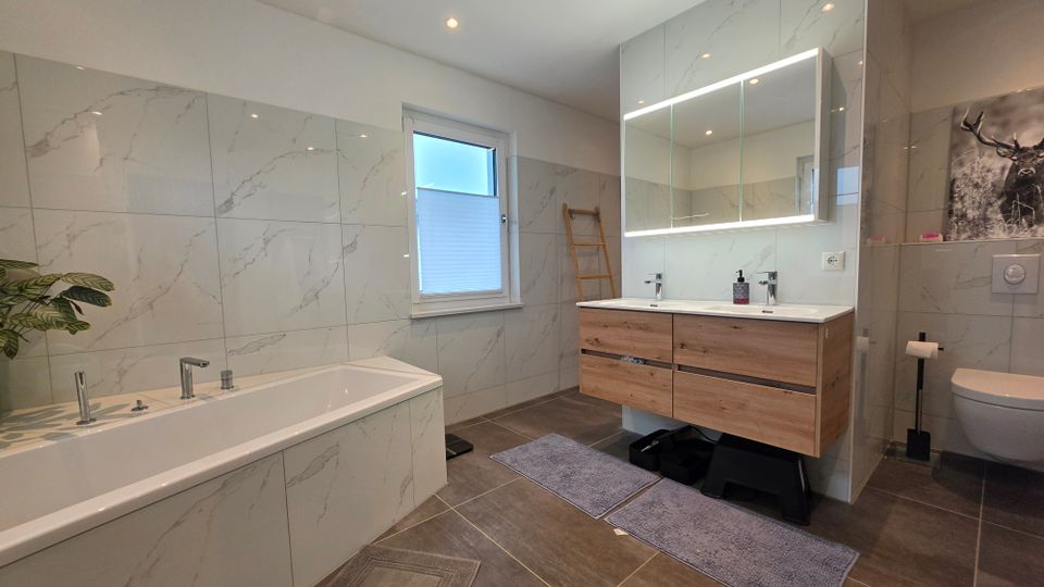 Modernes Badezimmer mit marmorierten Wandfliesen, großer Badewanne links und Doppelwaschtisch mit Holzunterschrank sowie beleuchtetem Spiegelschrank. Rechts ist eine wandhängende Toilette zu sehen; ein Fenster, Einbauspots und dunkle Bodenfliesen prägen den Raum.