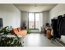 Ein modernes, kleines Zimmer mit schlichtem Interieur, bestehend aus einer orangefarbenen Couch, einem Schreibtisch mit Computer und Musikequipment, sowie einer Kleiderstange mit Kleidung. Ein großes Fenster mit weißen Vorhängen bietet Blick auf ein Backsteingebäude, während grüne Zimmerpflanzen den Raum dekorieren.