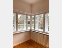 Ecke eines hellen Raumes mit großen, weißen Sprossenfenstern auf zwei Seiten, einem weißen Heizkörper unter den Fenstern und einem Boden aus Holzdielen. Draußen sind Bäume, Sträucher und ein Zaun sichtbar.