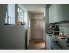 Schmale, moderne Küche mit glatten, matten Schränken in dezenter Farbe und einer Arbeitsfläche, die mit einer Kaffeemaschine und Lebensmitteln belegt ist. Ein kleines Fenster mit Dekorationsgegenständen lässt Licht ein, und im Hintergrund führt eine Tür zu einem weiteren Raum.