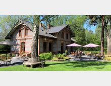 Historisches Backsteingebäude mit Zierelementen, umgeben von einem Garten mit grünen Bäumen und Rasenfläche; davor ein Außenbereich eines Cafés mit Holztischen, Stühlen und Schirmen.