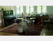 Ein Musikzimmer mit einem Schlagzeugset auf einem roten Teppich, einem schwarzen Klavier links und einem Schreibtisch mit Musikequipment sowie großen Fenstern im Hintergrund, die Licht in den Raum lassen.