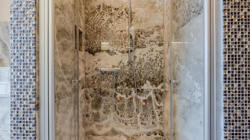 Moderne Dusche mit Glasabtrennung und Handbrause vor großformatigen, marmorierten Steinfliesen. Seitlich sind schmale Streifen aus kleinen Mosaikfliesen in Blau- und Brauntönen zu sehen.