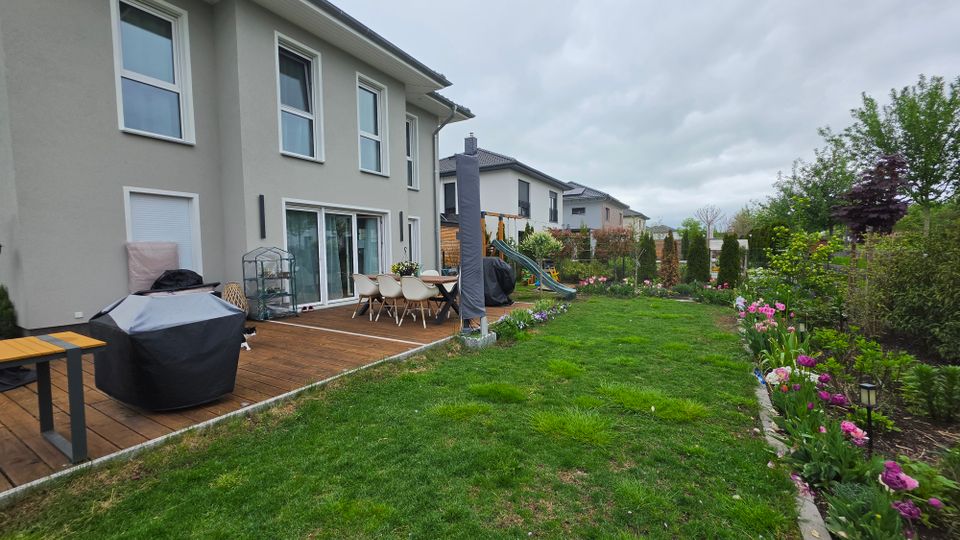 Blick in den Garten eines modernen Reihenhauses mit Holzterrasse, Esstisch und überdachten Gartenmöbeln. Daran schließt eine Rasenfläche mit Blumenbeeten, Sträuchern und Sichtschutzhecken an; im Hintergrund sind Nachbarhäuser und ein Spielgerät zu sehen.