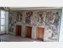 Wandmosaik in einem Gebäude mit drei hölzernen Türen. Das Mosaik zeigt verschiedene Alltagsszenen mit Menschen, darunter Kinder, Erwachsene bei Arbeit und Freizeit sowie symbolische Elemente wie Pflanzen, Tiere und Werkzeuge. Links befindet sich eine große Fensterfront mit buntem Glas.