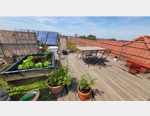 Dachterrasse mit Holzboden, bepflanzten Hochbeeten, Blumentöpfen und einem Tisch mit Klappstühlen; im Hintergrund sind geländerte Bereiche mit Begrünung und Solarzellen sowie Ziegeldächer zu sehen.