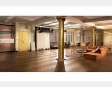 Weitläufiger Loftbereich mit hölzernem Fischgrätenparkett, gelb gestrichenen Metallsäulen und hohen Decken. Der Raum ist möbliert mit einer orangefarbenen Eckcouch, einem Glastisch, Stühlen und dezenten Beleuchtung; im Hintergrund stehen Requisiten und Fotostudioequipment.