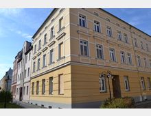 Eckansicht eines mehrstöckigen Wohngebäudes im klassischen Baustil mit gelber Fassade und weißen Fensterrahmen. Daneben befinden sich ähnliche Gebäude in verschiedenen Pastelltönen, und im Vordergrund steht eine Straßenlaterne an einem Gehweg mit kleinen Grünflächen.