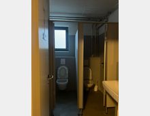 Zweikabinen-Toilettenraum mit halbhohen Trennwänden, einem kleinen Fenster in der Rückwand und sichtbaren Rohrleitungen an der Decke. Rechts im Vordergrund befindet sich ein Waschbecken.