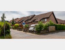 Reihenhaussiedlung mit rotbraunen Klinkerfassaden und steilen Satteldächern, umgeben von Gärten mit Zäunen und Hecken. Im Vordergrund verläuft eine schmale Wohnstraße mit Straßenverkehrsschildern und einem parkenden Wohnmobil.