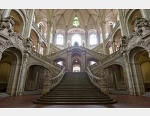 Großes historisches Treppenhaus mit kunstvoll verzierten Balustraden und steinernen Skulpturen in einem Gebäude mit hohen, spitzbogigen Decken und großen Fenstern, durch die Tageslicht einfällt. Die zentrale breite Treppe teilt sich in zwei seitliche Aufgänge, die von Bögen und Säulen eingerahmt sind.