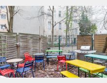 Kleiner, eingezäunter Außenbereich mit mehreren bunten Tischen und Stühlen, Holzsichtschutzwänden, einem Grill und Bäumen. Im Hintergrund sind Wohngebäude und ein Spielplatz zu sehen.