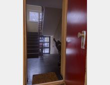 Blick aus einer geöffneten roten Tür in ein Treppenhaus eines Wohnhauses mit schwarzen Stufen, einem quadratischen Fenster, einer Glastür im Erdgeschoss und einem Kinderwagen an der Wand. Der Boden ist mit dunklen Gummiflachnoppen bedeckt, und eine Fußmatte liegt vor der Tür.