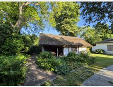 Reetgedecktes Haus in einem Garten mit üppiger Vegetation, umgeben von Bäumen und Büschen. Ein gepflasterter Weg führt zum Eingang, daneben befinden sich Blumenbeete und Grünflächen.