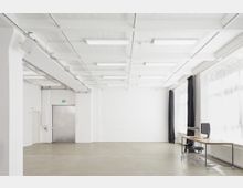 Minimalistischer, heller Raum mit weißen Wänden, Sichtbetondecke und großflächigen Fenstern auf der rechten Seite, teils mit schwarzen Vorhängen. Links eine Metalltür mit Notausgangsschild, rechts ein einfacher Schreibtisch mit Computer und schwarzem Bürostuhl auf Rollen.