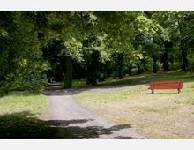 Parklandschaft mit einem geschwungenen Kiesweg, umgeben von Bäumen mit dichtem Laub. Auf einer Rasenfläche steht eine rote Parkbank rechts vom Weg.