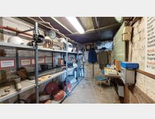 Ein kleiner Raum mit Regalen voller Ausrüstungsgegenstände, darunter Helme, Feuerlöscher und Schläuche, vermutlich aus einem historischen Feuerwehr- oder Zivilschutzkontext. An der Wand hängen Uniformen, und ein Tisch mit alter Büroausrüstung steht auf der rechten Seite.