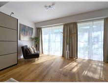 Helles Wohnzimmer mit Holzboden, bodentiefen Fenstern mit weißen Vorhängen und beigen Gardinen. Ein schwarzer Sessel mit Kissen, ein abstraktes, buntes Gemälde an der Wand und eine große weiße Vase mit Gräsern ergänzen die Einrichtung.