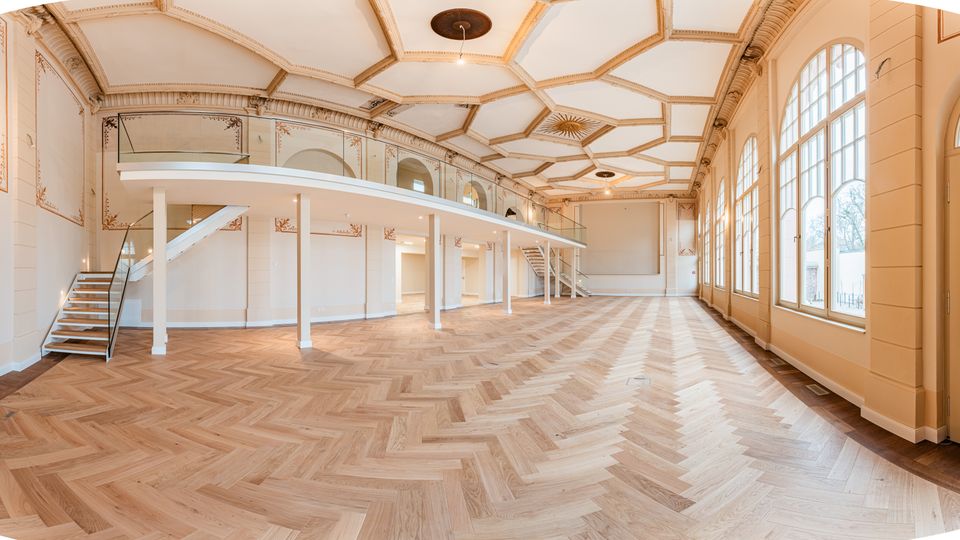 Großer, leerer Saal mit hellem Fischgrät-Parkett, hoher kassettierter Decke mit Stuck und umlaufender Empore mit Treppen. Rechts sorgen bodentiefe, bogenförmige Fensterfronten und eine Glastür für viel Tageslicht.