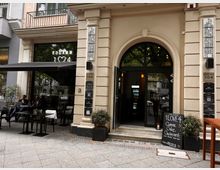 Außenansicht eines Cafés in einem historischen Gebäude mit cremefarbener Fassade und einem großen, bogenförmigen Eingang mit Glastür. Vor dem Café stehen schwarze Tische und Stühle unter einer markanten schwarzen Markise, mit ein paar Gästen, die dort sitzen.