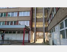 Innenhof zwischen zwei mehrstöckigen Betongebäuden mit langen Fensterbändern und einem überdachten Eingang mit Stützen. In der Mitte führt ein Durchgang unter einem verbindenden Gebäudeteil nach außen, flankiert von hohen Betonpfeilern und gepflastertem Boden.