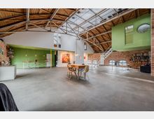 Großer, offener Loft‑Innenraum in einer umgebauten Industriehalle mit Sichtziegelwänden, Betonboden und hoher Holzdachkonstruktion mit Stahlträgern und Oberlichtern. In der Mitte stehen Tisch und Stühle, rechts führt eine Metalltreppe zu einem grün gestrichenen Zwischengeschoss, daneben Regale und Sitzbereich.