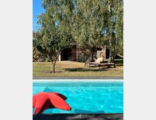 Ein Außenbereich mit Swimmingpool im Vordergrund, in dem ein rotes aufblasbares Objekt schwimmt. Im Hintergrund stehen Bäume und ein Haus mit einer Fassade aus Stein sowie Liegestühle auf einer Rasenfläche.