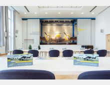 Innenraum eines Konferenzraums mit einer großen Wandgestaltung eines historischen Flugzeugs im Hangar im Hintergrund und dem Schriftzug „Flugplatz Schönhagen“. Im Vordergrund stehen Konferenztische mit blauen Stühlen, auf denen Broschüren verteilt sind.