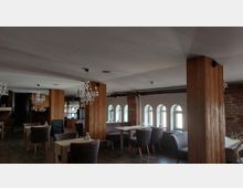 Ein gemütliches Restaurant mit Holztischen, gepolsterten Stühlen und großen Holzsäulen. An der Decke hängen Kristallleuchter, und an der Wand sind schmale Fenster mit Rundbögen, die Tageslicht hereinlassen.