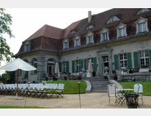 Großes historisches Gebäude mit rotem Ziegeldach und mehreren Dachgauben, grünen Fensterläden und einer Terrasse mit Sitzmöbeln. Im Vordergrund befinden sich weiße Klappstühle, Sonnenschirme und ein gepflasterter Weg, der die gepflegte Rasenfläche umrahmt.