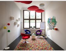 Kleiner Besprechungsraum mit mehreren bunten Design-Stühlen, einem runden Tisch in der Mitte und einer Staffelei mit Flipchart. Die Wände sind mit kreativen Elementen wie Wandkugel-Lampen und einer bunten Lego-ähnlichen Installation dekoriert, unter einem großen Fenster mit Gittermustern.