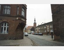 Straßenszene in einer Altstadt mit Kopfsteinpflaster, flankiert von Ziegelbauten. Im Hintergrund erhebt sich eine Kirche mit spitzem Turm, umgeben von Wohngebäuden aus mehreren Epochen.