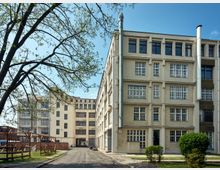 Historischer Industriekomplex mit vierstöckigen Gebäuden aus hellem Putz und großen, gitterförmigen Fenstern; eine gepflasterte Straße führt durch die Anlage, umgeben von niedrigem Grün und vereinzelten Bäumen.