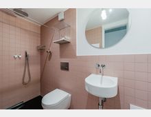 Kleines Badezimmer mit rosa Wandfliesen, bodengleicher Dusche mit Regenduschkopf und Handbrause sowie wandhängendem WC. Rechts sind ein kleines Waschbecken mit Wandarmatur und ein runder Spiegel über der halbhoch gefliesten Wand zu sehen.